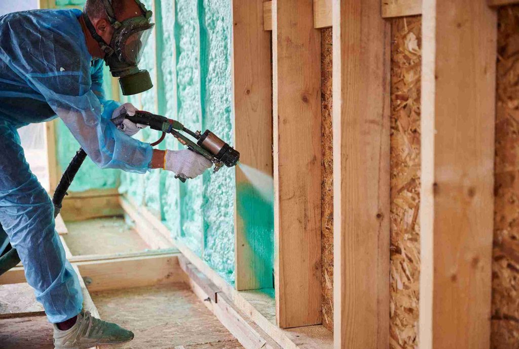 A man in a blue suit applies foam insulation with a spray gun, focusing on a wall in a construction setting.